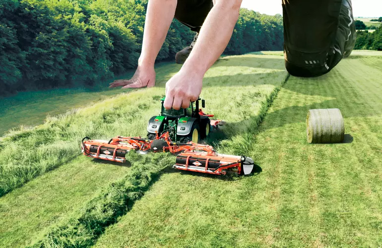 Eine Darstellung des FC 9330 RA, einer Dreifach-Scheiben-Mähknickzetter-Kombination der Firma KUHN, die wie ein Spielzeug von einem großen Menschen bewegt wird.