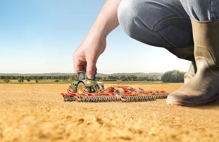 Eine Darstellung des OPTIMER 12000 L, einer Kurzscheibenegge der Firma KUHN, die wie ein Spielzeug von einem großen Menschen bewegt wird.