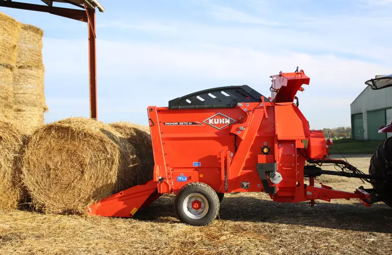 Ein Bild des KUHN PRIMOR 3570 M - einem Einstreu- und Verteilgerät - welches mit einem Rundballen beladen wird., welche durch einen DNA-Strang verbunden sind.