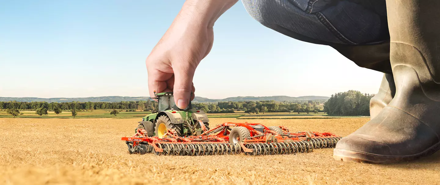 Eine Darstellung des OPTIMER 12000 L, einer Kurzscheibenegge der Firma KUHN, die wie ein Spielzeug von einem großen Menschen bewegt wird.