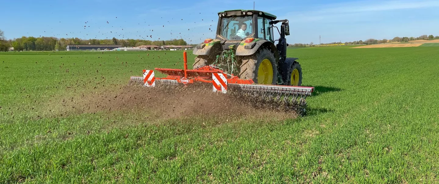 Un tracteur tracte la houe rotative STARLINER rouge de KUHN dans un champ verdoyant, travaillant le sol en bandes. La scène illustre un chantier agricole sous un ciel bleu dégagé, avec des oiseaux visibles dans les airs.